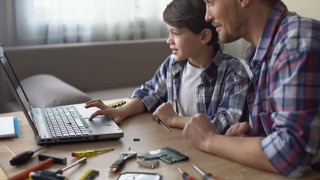 Son and father sitting at table, searching PC video tutorial on laptop together