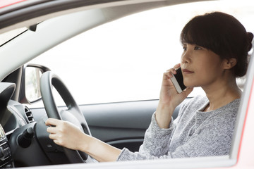 Young asian woman in casual wear driving a car, using smart phone and  looking on road. Risky driving. Woman sitting in car with cell phone