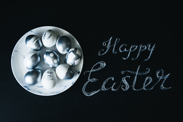 Group of diverse silver Easter eggs in a marble plate with inscription Happy Easter on black background, stylish original idea, selective focus