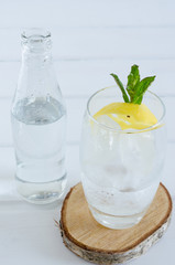 Sparkling water in glass cup with lemon and mint branch on white wooden background.