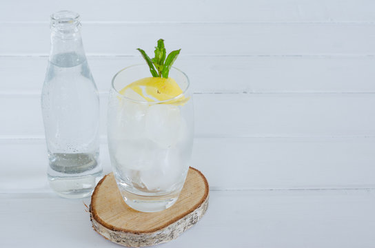 Sparkling water in glass cup with lemon and mint branch on white wooden background.