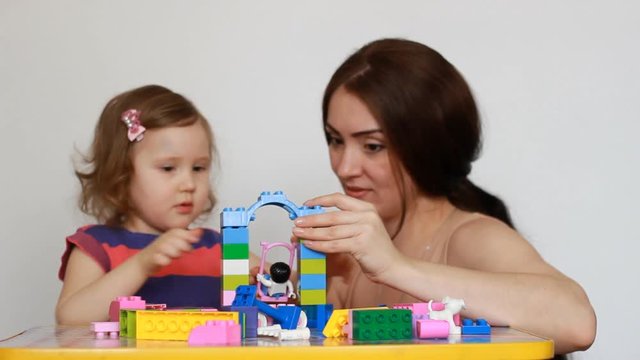 Mother and her little daughter playing together sitting at the table. Girl busy with his building constructor, mom watching and talking with child