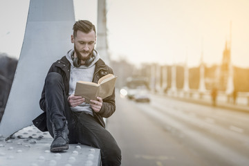 A retro style photo of a young hipster man sitting on a bridge and reading a book.