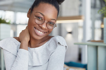 Pleased attractive dark skinned female model wears glasses, has shining smile, glad to finish work and has break, poses against office interior. Glad African American woman with happy expression