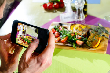 Man taking photo delicious fish served with tomatoes, lemon and herbs on the wooden board at a cafe. Selective focus