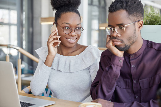 Two African American groupmates meet together for making project work or prepare for classes: happy dark skinned female speaks with friend via cell phone, sits near her parnet who is focused in laptop
