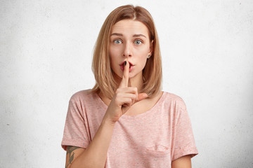 Beautiful young female model asks to be quiet people who surround her, looks with warm blue eyes at...