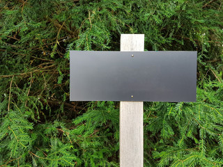 A blank black sign, nailed to a wooden post in front of a green hedge.