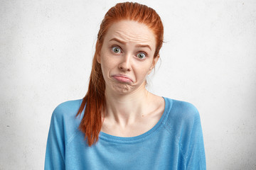 Beautiful clueless female has red hair, curves lips, looks with dissatiasfied expression, being discontent and puzzled, expresses uncertainty, poses on white concrete wall. People, facial expressions