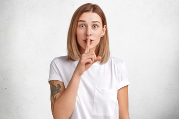 Pretty young female keeps fore finger on lips, demonstrates silence sign, has puzzled surpirsed...