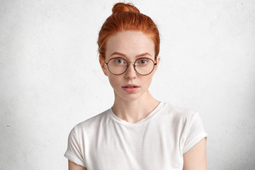 Photo of serious clever redhead female student with ginger hair knot, wears round spectacles, comes on lecture at university, isolated over white concrete background. Confident ginger woman in eyewear