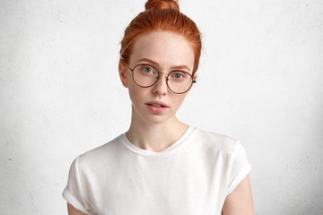 Fototapeta premium Horizontal portrait of serious redhaired female in big round spectacles looks with mysterious expression directly into camera, has freckled healthy skin, contemplates about how do important task