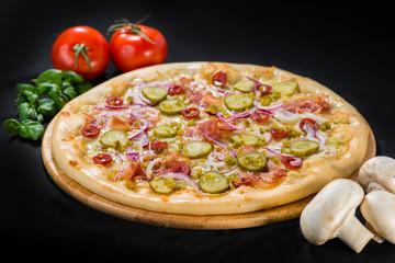 Pizza with mozzarella, hunting sausages, bacon, pickled cucumbers and pepper on wooden board on dark background