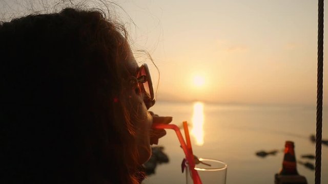 Young pretty woman in sunglasses enjoying a tropical tequila sunrise alcoholic cocktail decorated with flower at the seaside at amazing sunset on her summer vacation. slow motion. 1920x1080