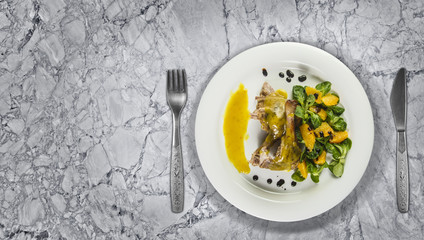 Duck legs with orange sauce and lamb's lettuce salad with oranges and vinegar on white plate on marble counter top.