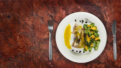 Duck legs with orange sauce and lamb's lettuce salad with oranges and vinegar on white plate on granite counter top.