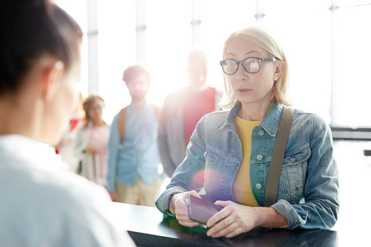 Senior woman in casualwear and eyeglasses looking at check-in manager while talking to her