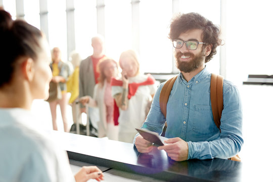 Happy young bearded man in eyeglasses and denim shirt showing his passport to check-in receptionist before flight