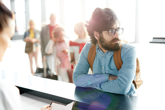 Bearded traveler with backpack leaning by counter while waiting for check-in of his documents before flight