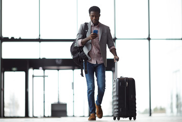 Young mobile traveler with smartphone, backpack and suitcase walking along airport and messaging or...