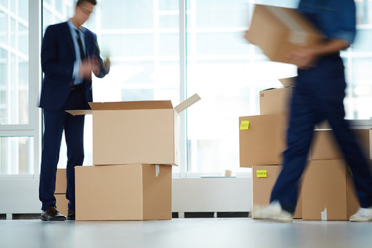 Businessman standing over open box by window of office center while relocation service worker carrying packages