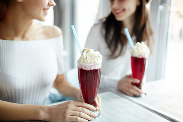 Young women sitting in cafe and having talk by glass of cocktail with whipped cream top
