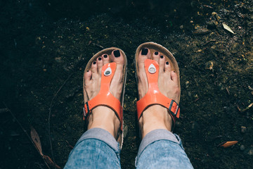 Woman's feet in orange sandals and jeans standing near the lake