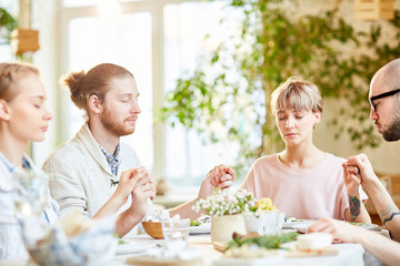 Four young people holding by hands while sitting by served table and saying pray before dinner