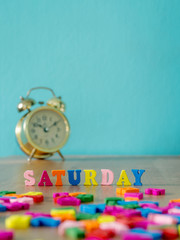 Colorful wooden word SATURDAY on wooden table and vintage alarm clock and background is powder blue. English alphabet made of wooden letter color.