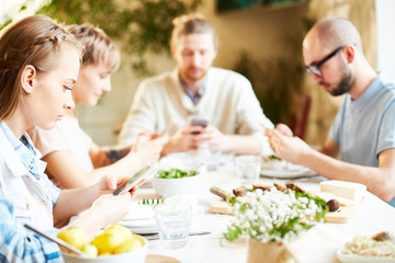 Group of young people searching or texting in their gadgets while sitting by served table by dinner