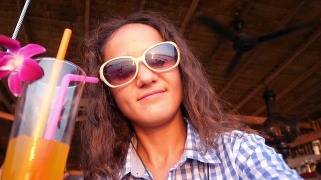 Attractive joyful girl in sunglasses enjoying tropical cocktail decorated with fresh flower and listen to music in headphones looks at sunset. slow motion. 1920x1080