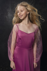 Obraz premium Beautiful curly blonde teenager child girl wearing a pink air dress made of veil swith stands in the wind which made flying her hair, and luxorioly smiles