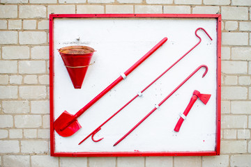 The fire shield is hanging on a brick wall. The old set of tools for protection includes a bucket, shovel, ax, scrap. Red firefighters are on a white background