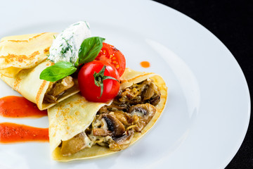 Homemade tasty pancakes (blinis) served with mushrooms and tomatoes in the white plate on dark background