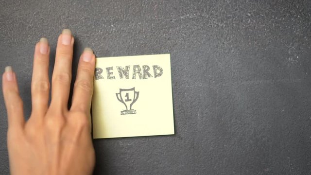 Closeup woman's hand sticking note with Reward word on the blackboard - video in slow motion