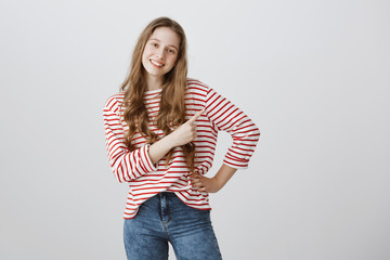 Follow me there. Studio shot of confident friendly teenage girl with blonde hair and positive smile in stylish striped sweater and jeans posing and pointing left with index finger over gray wall