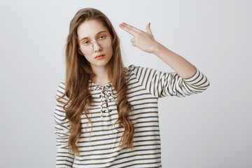 I would rather kill myself. Portrait of bored and fed up young woman in glasses making gun gesture and mimicking as if committing suicide, expressing annoyance and boredom, standing over gray wall