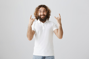 Your idea rocks. Studio shot of excited and joyful adult eastern man with beard showing rock signs and gazing at camera with widened eyes, smiling broadly, being happy and amazed over gray wall