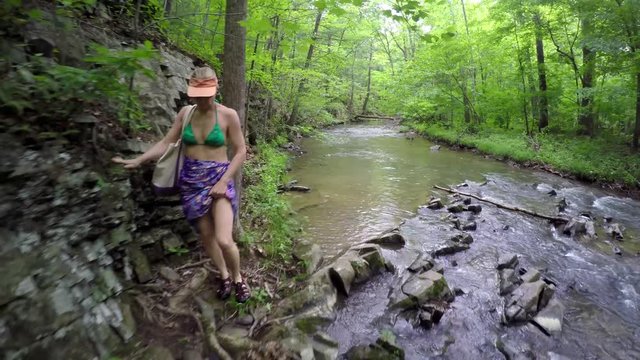 Bikini clad mature woman hiking through natural mountain stream or creek in forest, captured with steadicam