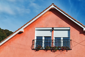 Attic windows of red stone house decorated with beautiful bright red flowers under blue sky. Gable roof and dormer type of roof outdoors.