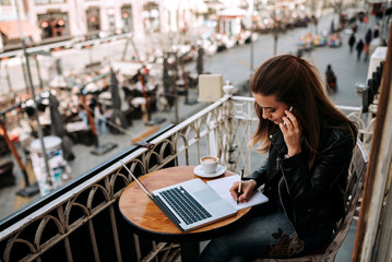 Young female entrepreneur working outdoors. Writting in a notebook while talking on a phone.