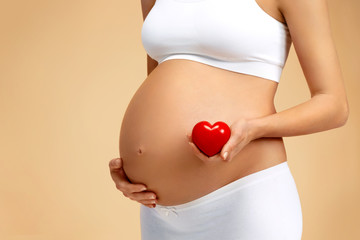 Close up of pregnant woman holding red heart on beige background. Pregnancy, maternity, preparation and expectation concept