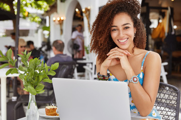 Photo of glad adorable curly African woman sits in front of opened laptop computer in sidewalk cafe, satisfied to make good presentation, has broad smile. Cheerful lovely young dark skinned woman