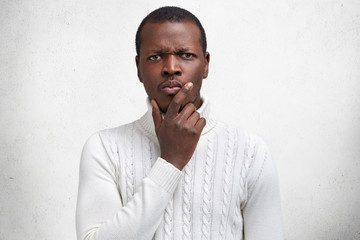 Waist up portrait of sullen serious young African American male looks with thoughtful gloomy expression, being dissatisfied with something, wears white sweater, isolated over studio concrete wall.