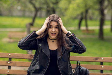 Glamorous young Caucasian woman in black leather jacket sitting in the park on the bench