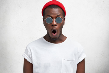 Stylish hipster guy wears trendy shades, white t shirt and red hat, keeps mouth rounded, being...
