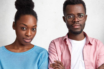 Indoor horizontal shot of confident attractive African American male and female looks with serious expression at camera, meet together to discuss future plans, isolated over white concrete wall