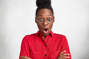 Beautiful emotional dark skinned businesswoman wears red formal blouse and spectacles, stares at camera with shocked expression, has deadline to prepare business project. Omg and ethnicity concept