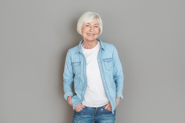 Senior woman in jeans jacket studio isolated on grey wall hands in pockets joyful