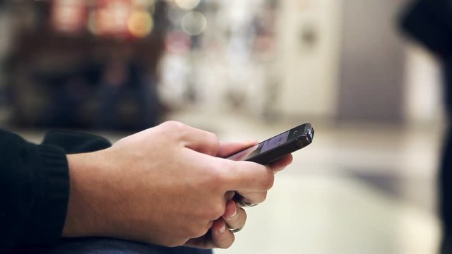 An impressive closeup of young man`s hands keeping a smartphone and scrolling the information while sitting in an airy shopping mall